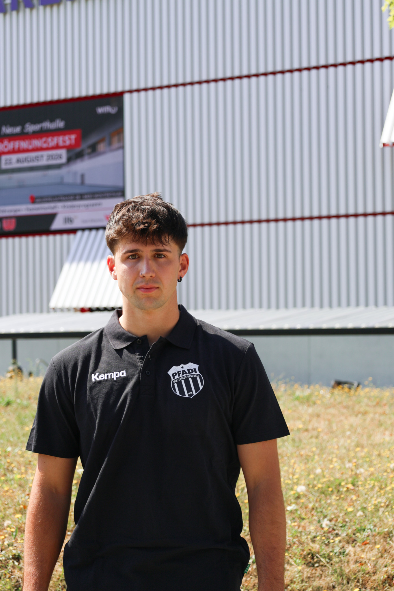 Junger Mann im schwarzen Kempa-Poloshirt mit Pfadi-Logo vor einer Sporthalle mit Eröffnungsfest-Banner.