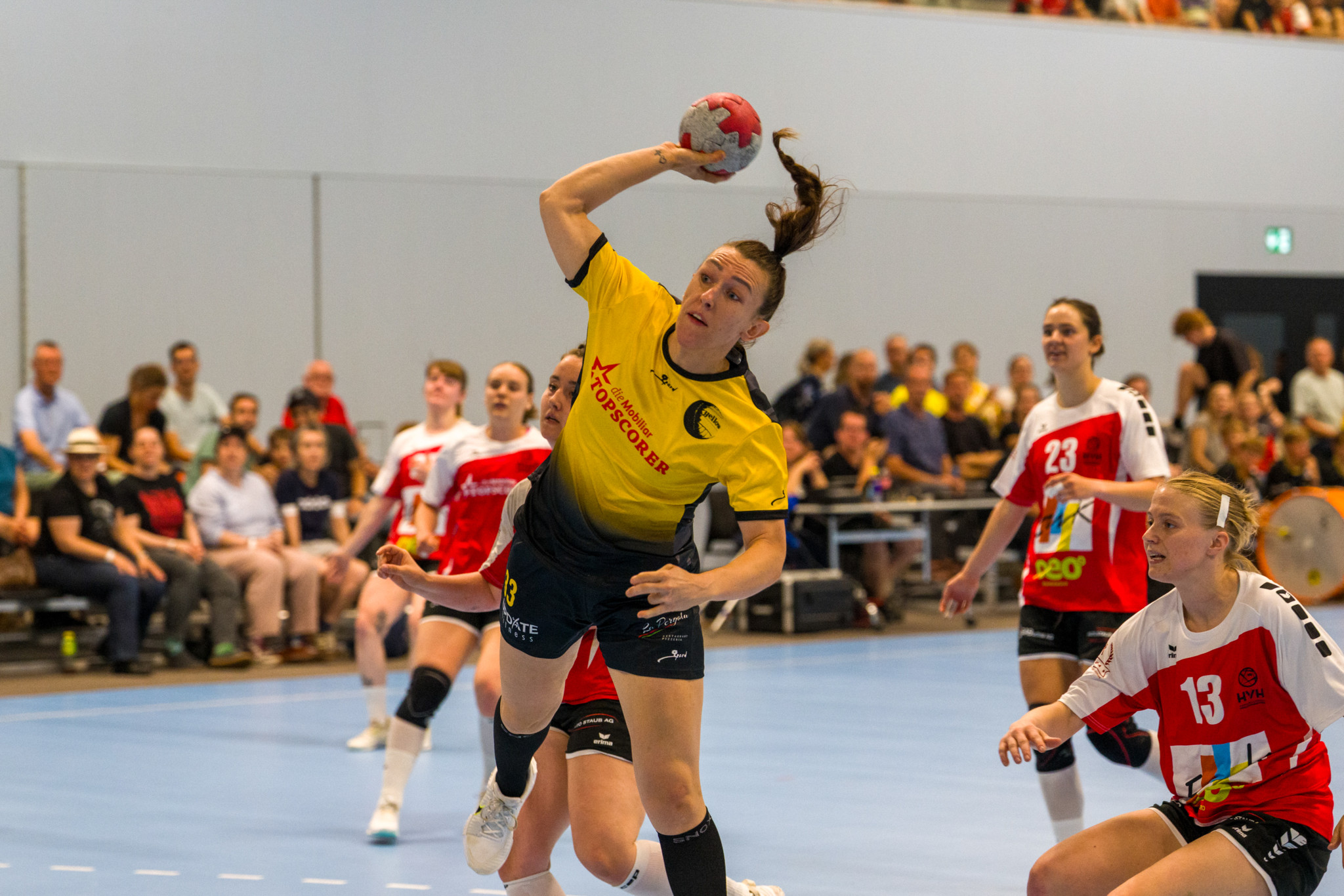 Handballspielerin im gelben Trikot springt hoch und wirft den Ball, umgeben von Gegnerinnen in roten Trikots in einer Sporthalle.