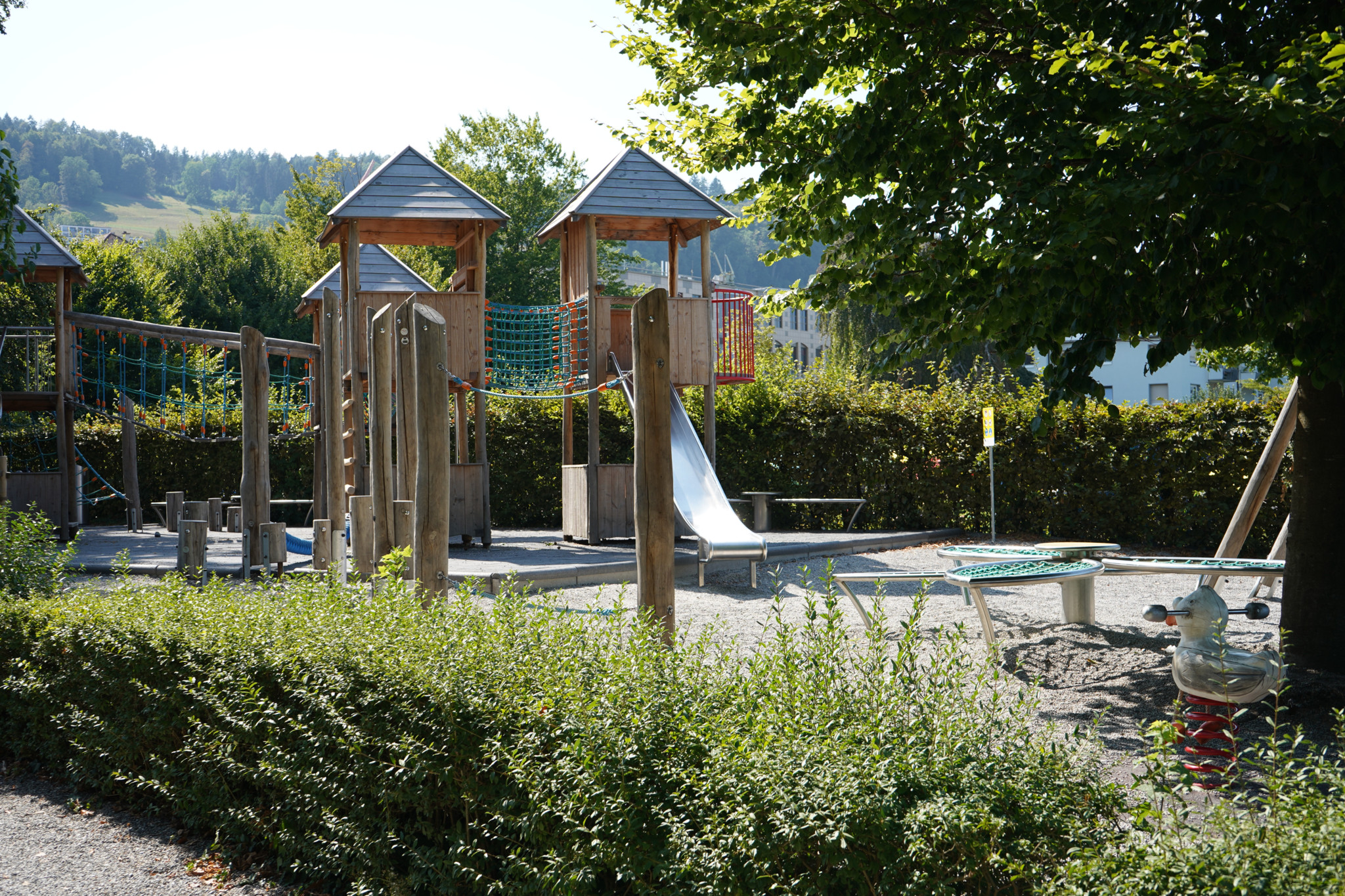 Kinderspielplatz Villa Seerose in Horgen mit Holzkletterturm, Rutsche und Sandkasten, umgeben von Büschen und Bäumen in ländlicher Umgebung.