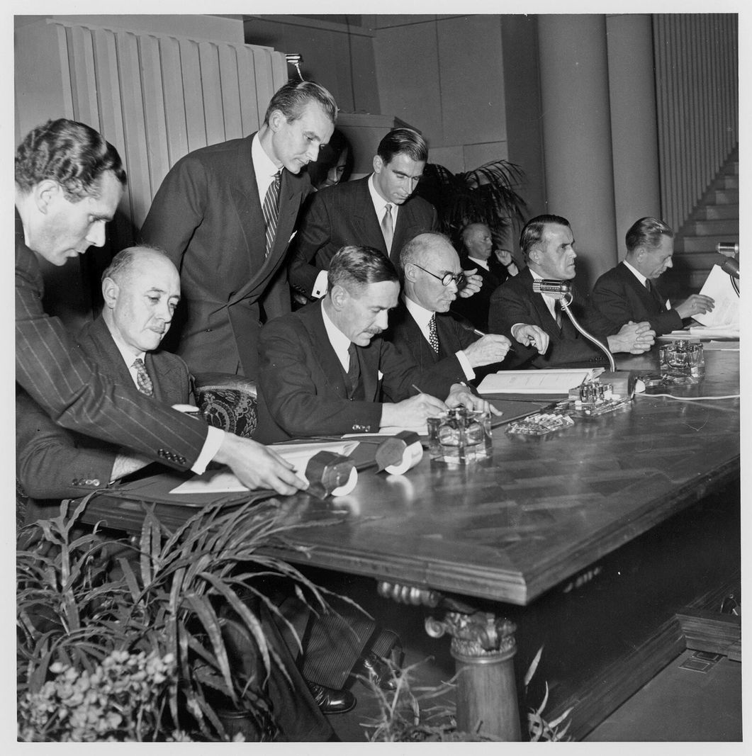 Des hommes en costume assis à une longue table signent des documents tandis que d'autres hommes debout les entourent.