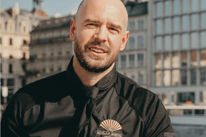 Chef en uniforme noir avec logo de Mandarin Oriental, souriant devant un bâtiment urbain.
