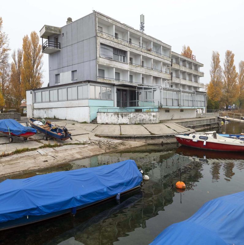Bâtiment gris abandonné près d’un quai avec des bateaux couverts sur l’eau, entouré d’arbres en automne.