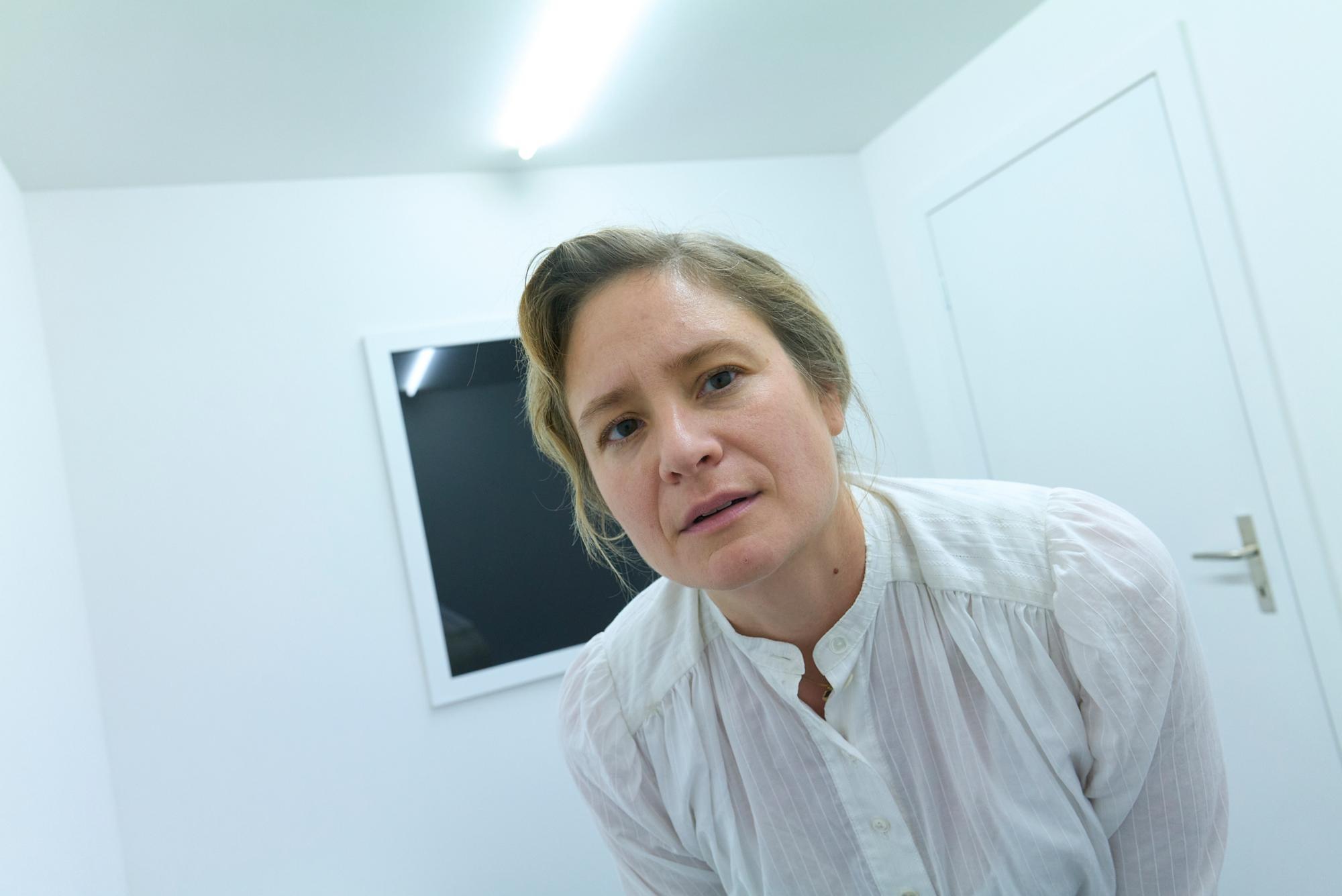 Frau in einem weissen Raum mit einem dunklen Bildschirm an der Wand, gekleidet in ein weisses Hemd.