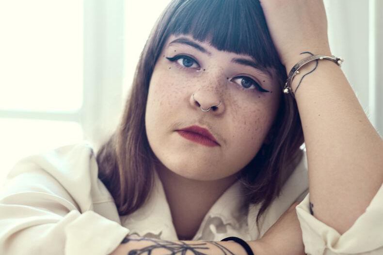 Femme aux yeux maquillés et cheveux bruns, portant une chemise blanche, pose avec une main sur la tête. Tatouage visible sur le bras.