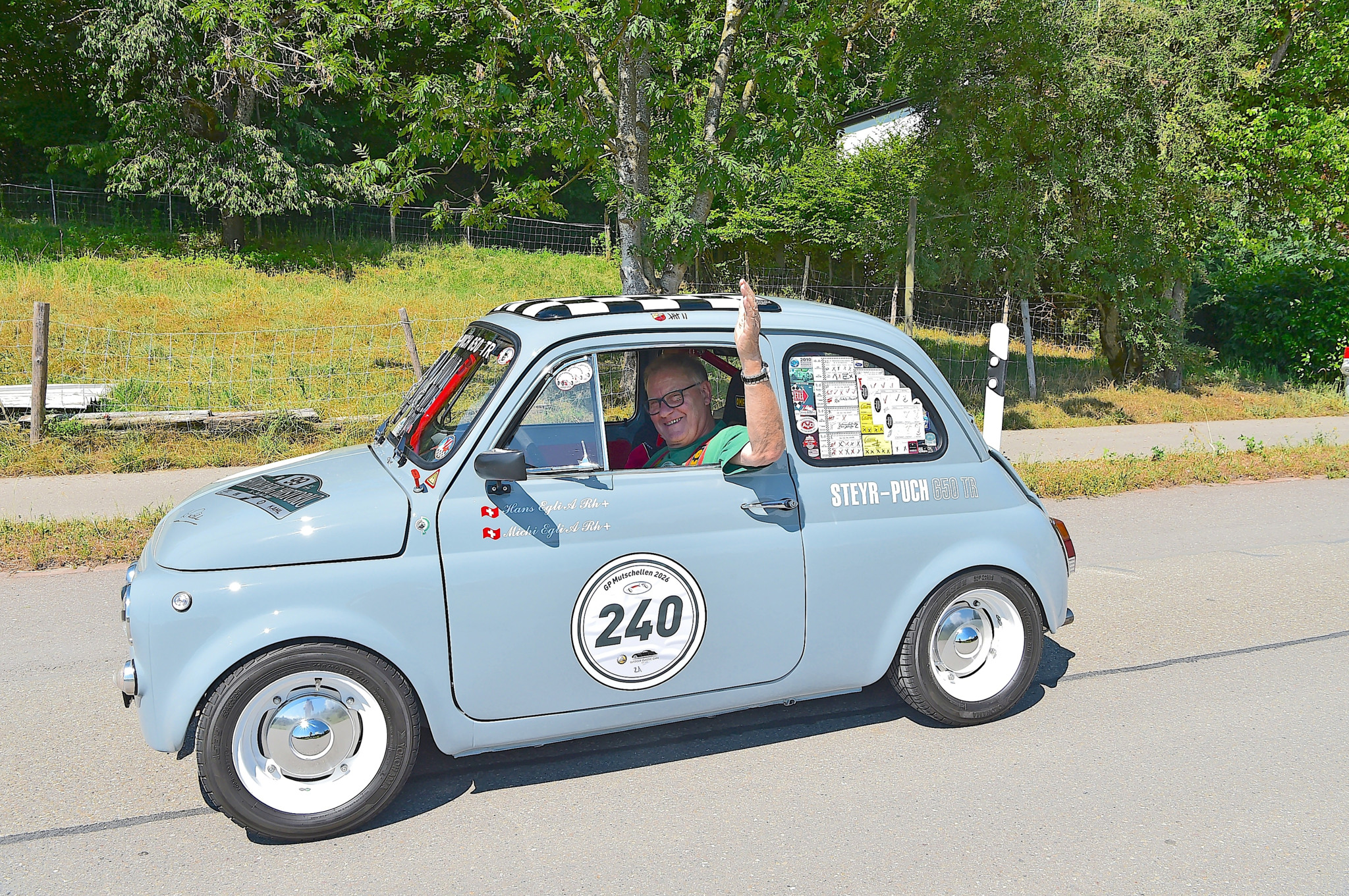 Fahrer winkt aus einem hellblauen Steyr-Puch Oldtimer mit der Startnummer 240 bei einer Oldtimerausfahrt.