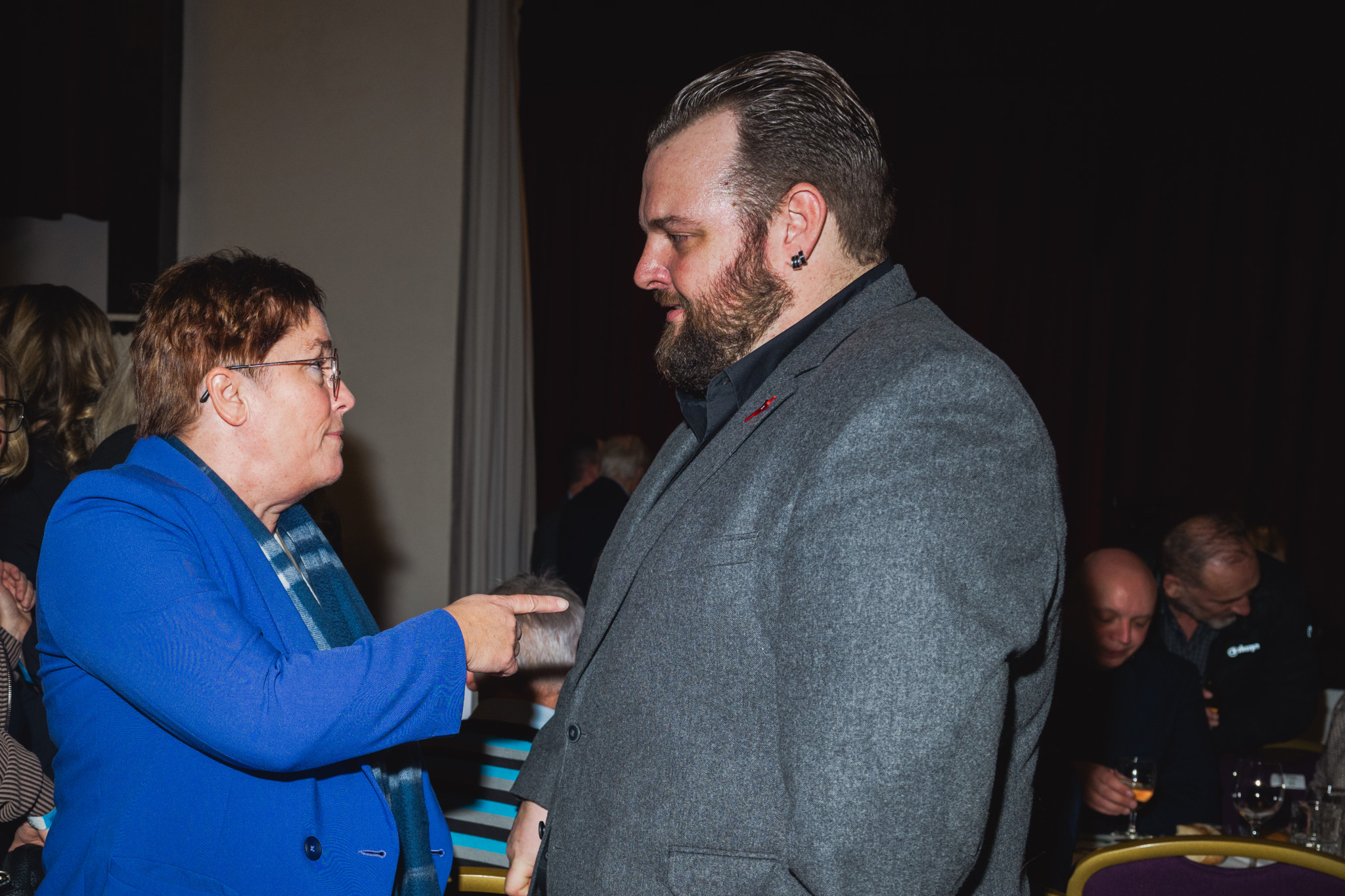 Eine ältere Frau im blauen Schal spricht mit einem Mann in einem grauen Anzug in einem Veranstaltungsraum.