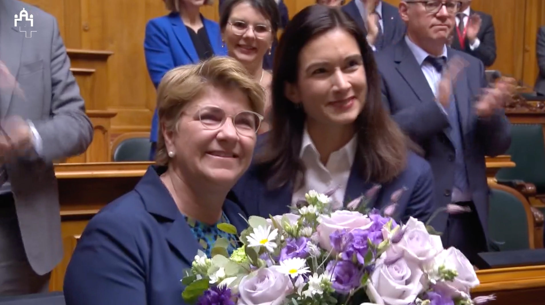 Deux femmes souriantes tenant un bouquet de fleurs mauves dans une salle de réunion parlementaire, entourées de personnes applaudissant.