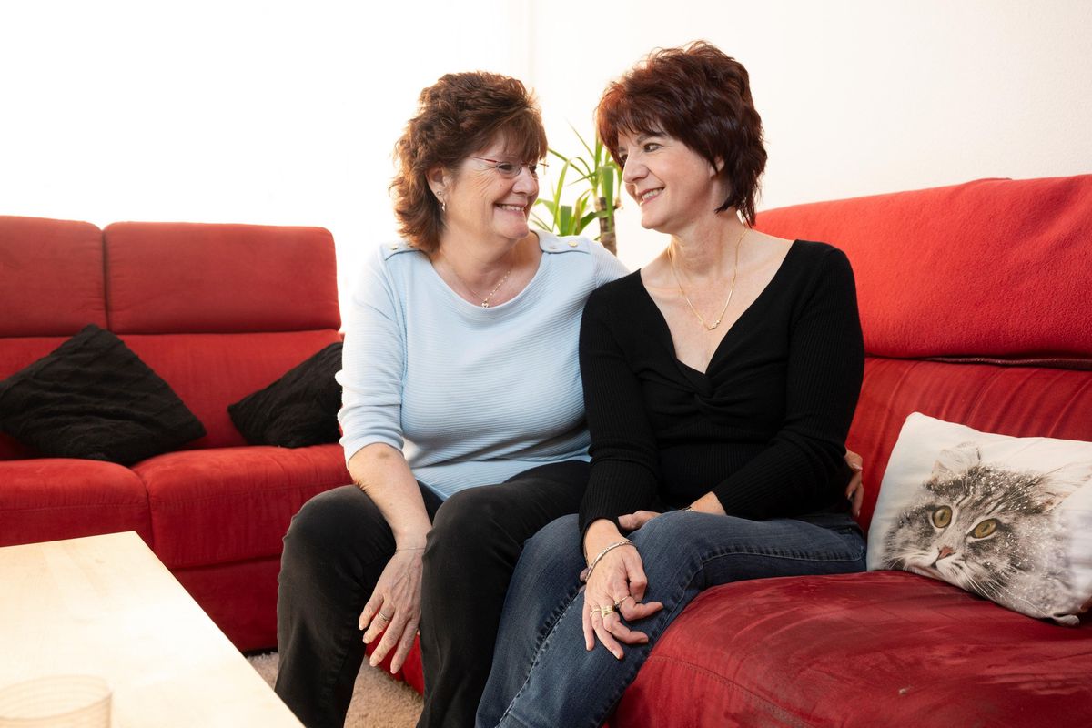 Deux sœurs assises sur un canapé rouge, Janique Baud en chemisier bleu clair et Marinette Baud en chemisier foncé, souriantes et proches.