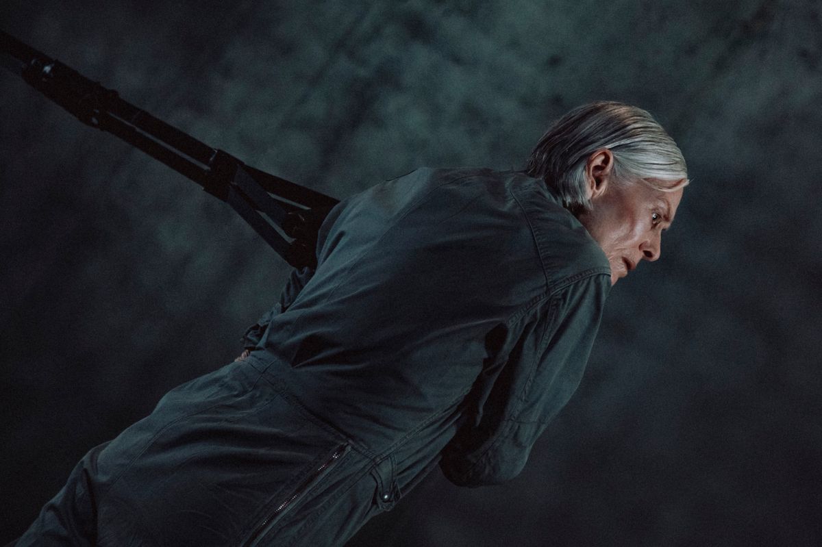 Personne âgée aux cheveux gris et courts, vêtue d'un manteau sombre, se tenant penchée vers l'avant avec un arrière-plan sombre.