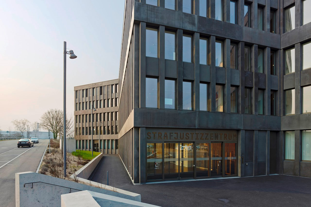 Modernes Strafjustizzentrum-Gebäude aus dunklem Beton mit verglasten Fassaden und beleuchtetem Eingangsbereich. Modernes Strafjustizzentrum-Gebäude aus dunklem Beton mit verglasten Fassaden und beleuchtetem Eingangsbereich.