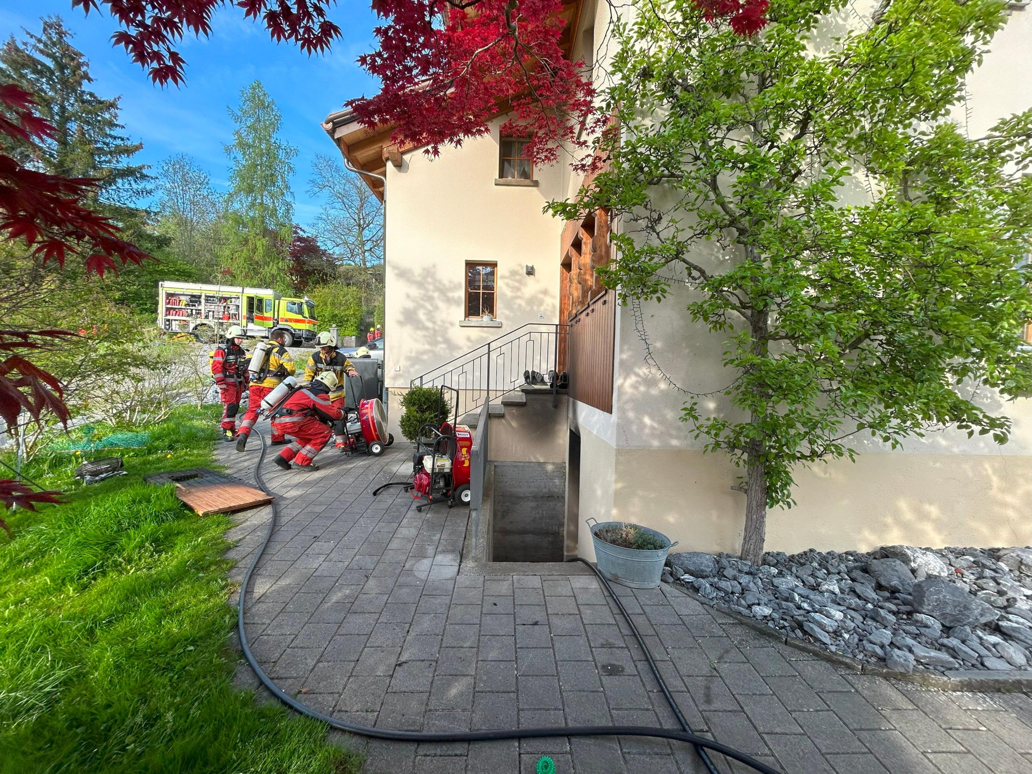 Feuerwehrleute in Schutzkleidung vor einem Wohnhaus, bereiten Schläuche und Ausrüstung vor. Ein Feuerwehrfahrzeug im Hintergrund.