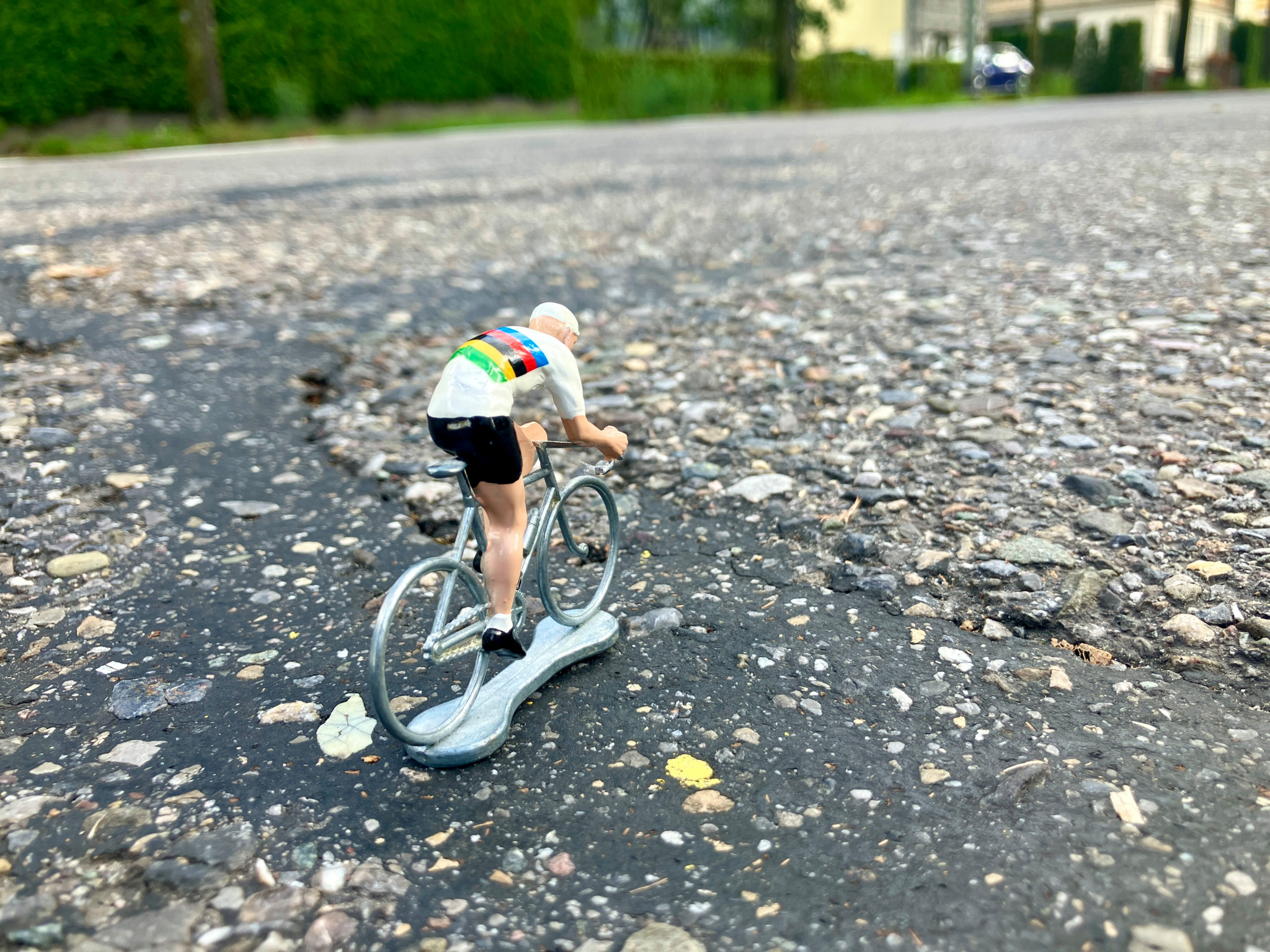 Detailaufnahme einer Miniaturfigur, die ein Trikot mit Regenbogenstreifen trägt und ein Fahrrad auf einer rauen Asphaltoberfläche fährt.
