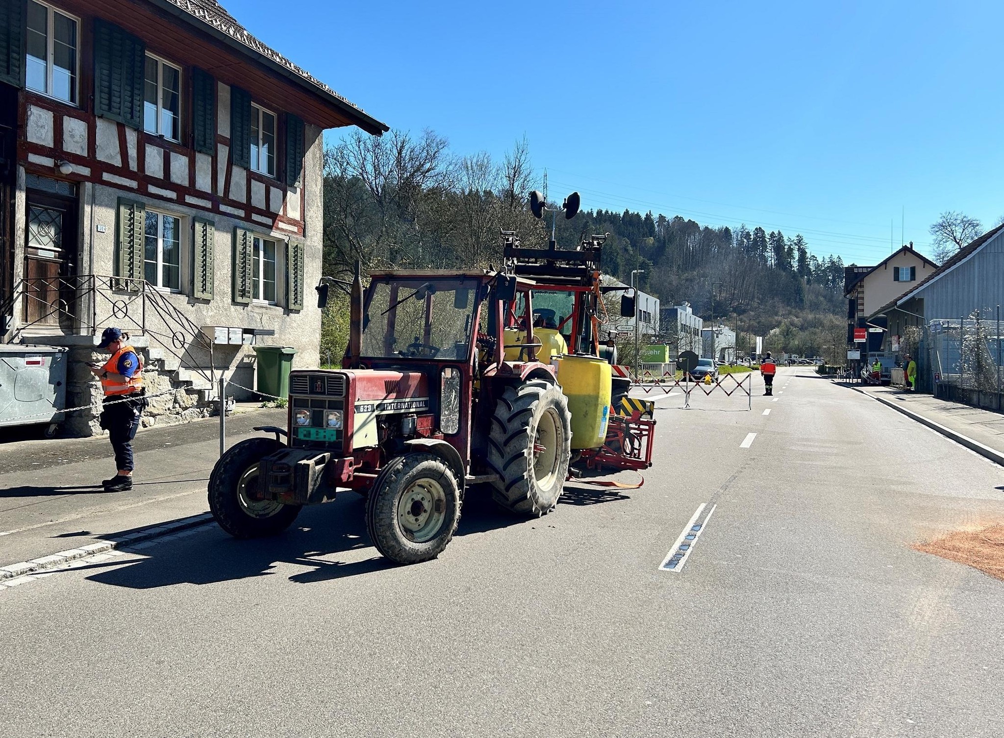 Ein Traktor mit Sprühausrüstung fährt auf einer leeren Strasse in einem Dorf. Rechts stehen zwei Personen in Schutzkleidung.