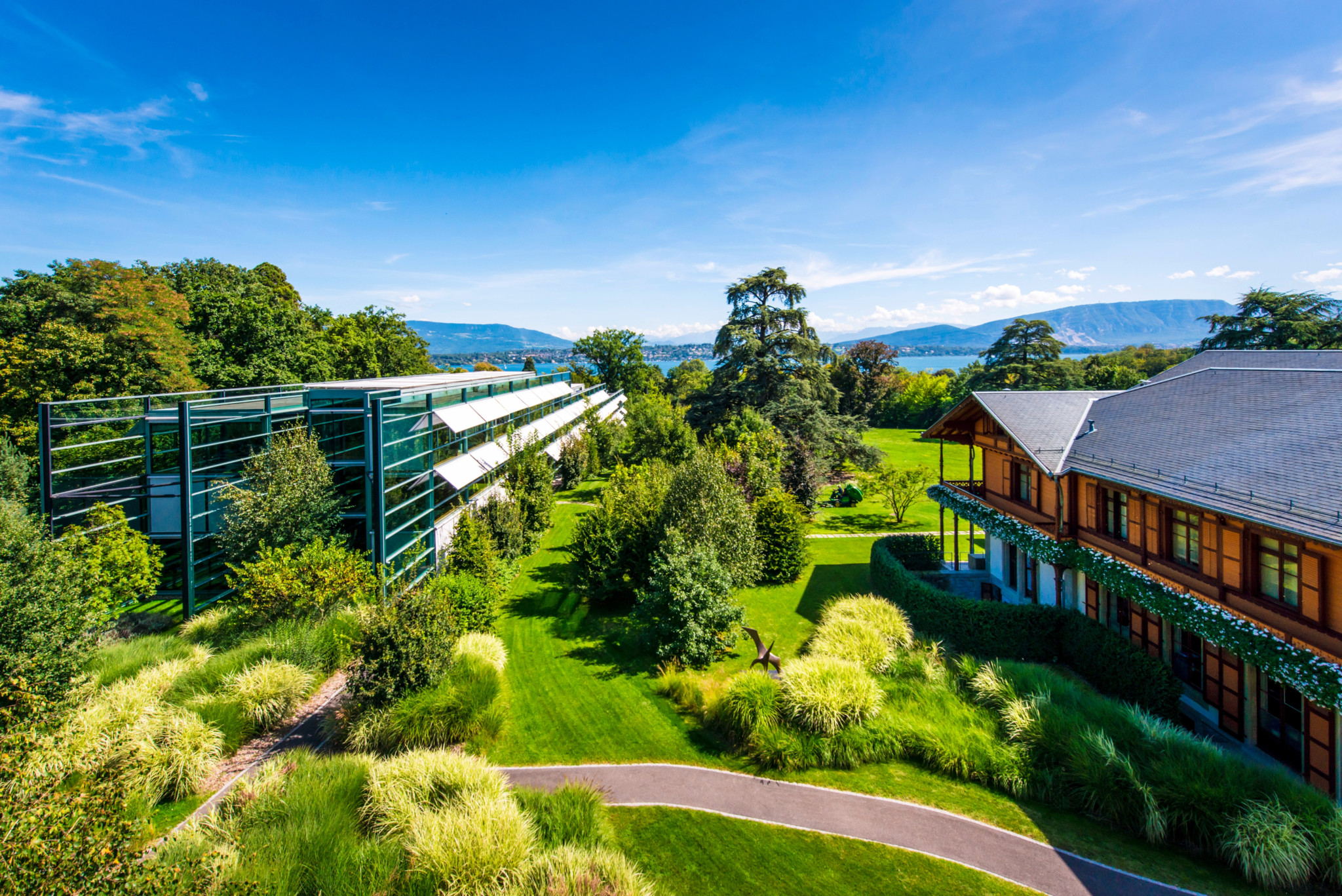 Gepflegte Gartenlandschaft mit modernen GlasgebÃ¤uden und traditionellem HolzgebÃ¤ude im Hintergrund, umgeben von grÃ¼nen BÃ¤umen.