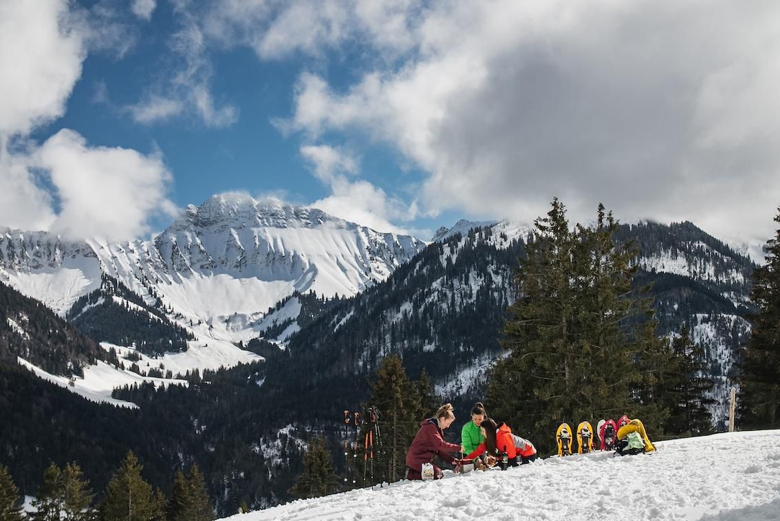 Paysage alpin enneigé avec un groupe de personnes se reposant, entouré de montagnes et de sapins, sous un ciel partiellement nuageux.