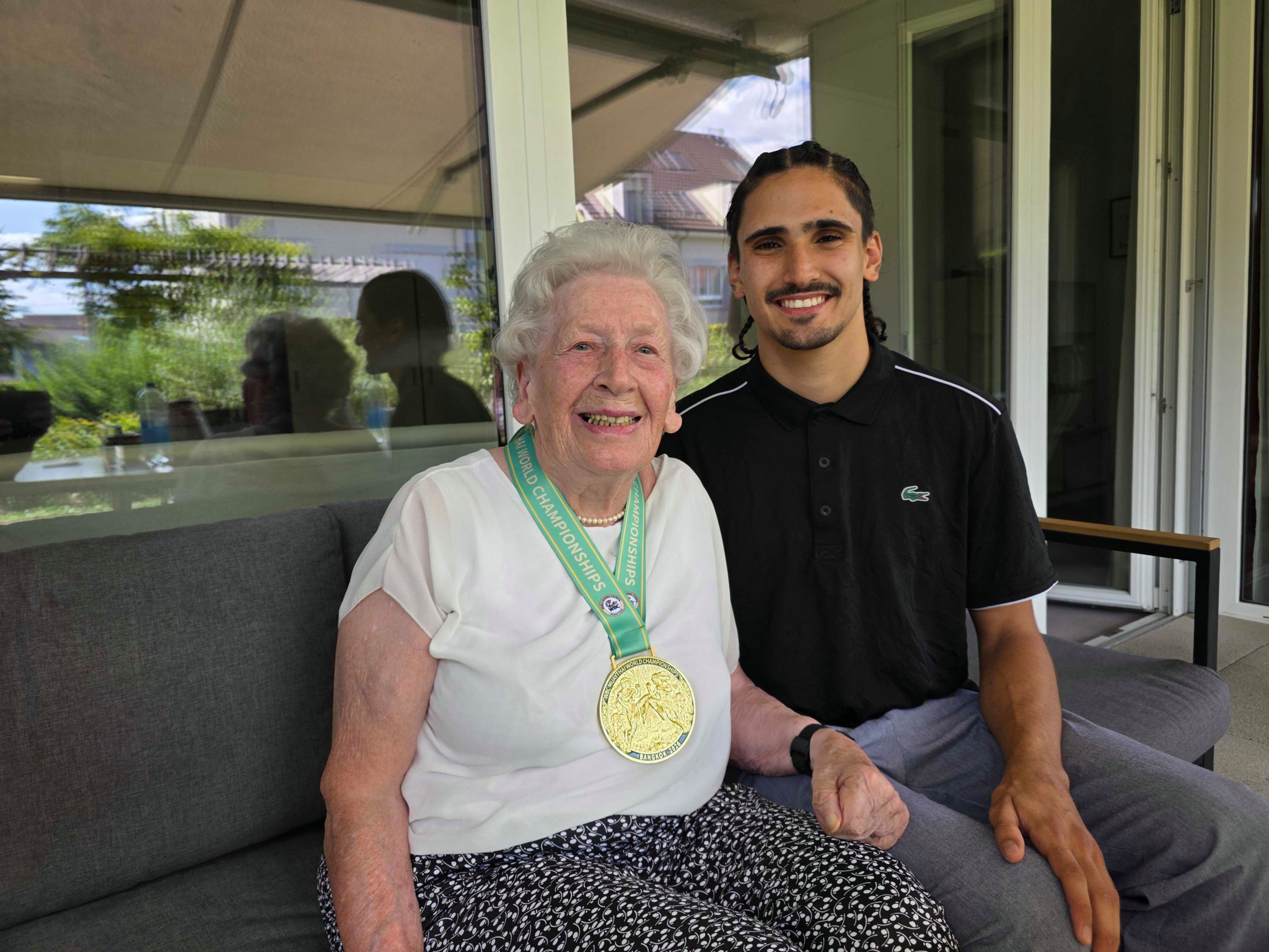 Ältere Frau mit goldener Medaille und grünem Band sitzt neben jungem Mann mit Bart auf einem Sofa im Freien.