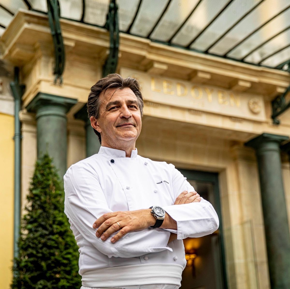 Chef en tenue blanche se tenant devant un bâtiment élégant avec des colonnes et un auvent en verre.