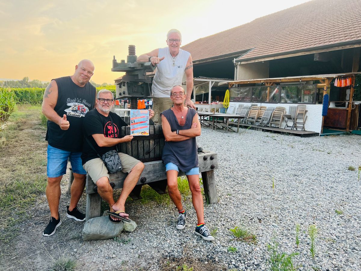 Quatre hommes souriants posant devant un bâtiment dans un cadre rural, certains assis et d'autres debout, lors d'une soirée d'été.