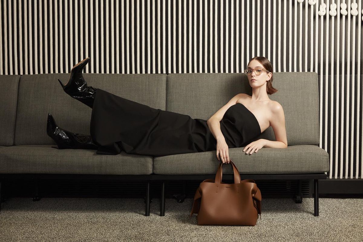 Femme allongée sur un canapé gris, portant une robe noire et des bottes noires, tenant un sac marron devant un mur rayé.