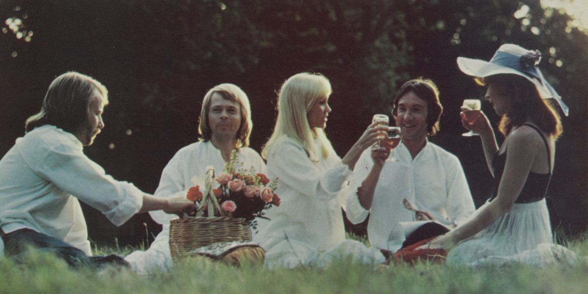 Un groupe de cinq personnes, habillées en blanc, pique-nique dans un parc avec un panier de fleurs et des verres à la main.