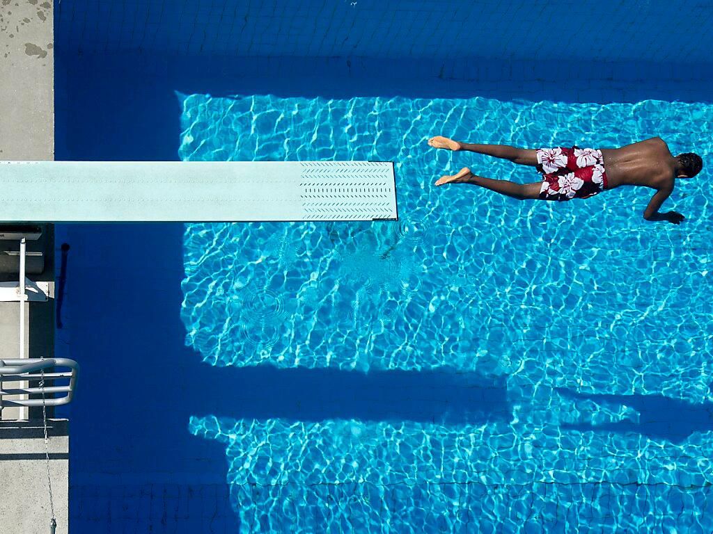 Vue aérienne d’une piscine extérieure avec un plongeoir. Une personne nage sous l’eau en portant un maillot de bain à fleurs.