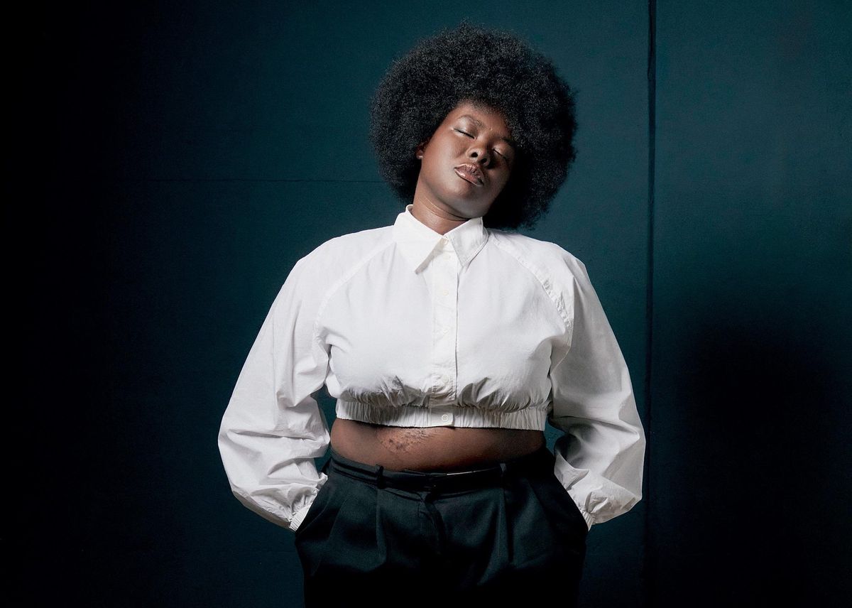 Personne portant une chemise blanche et un pantalon noir, posant devant un fond sombre.