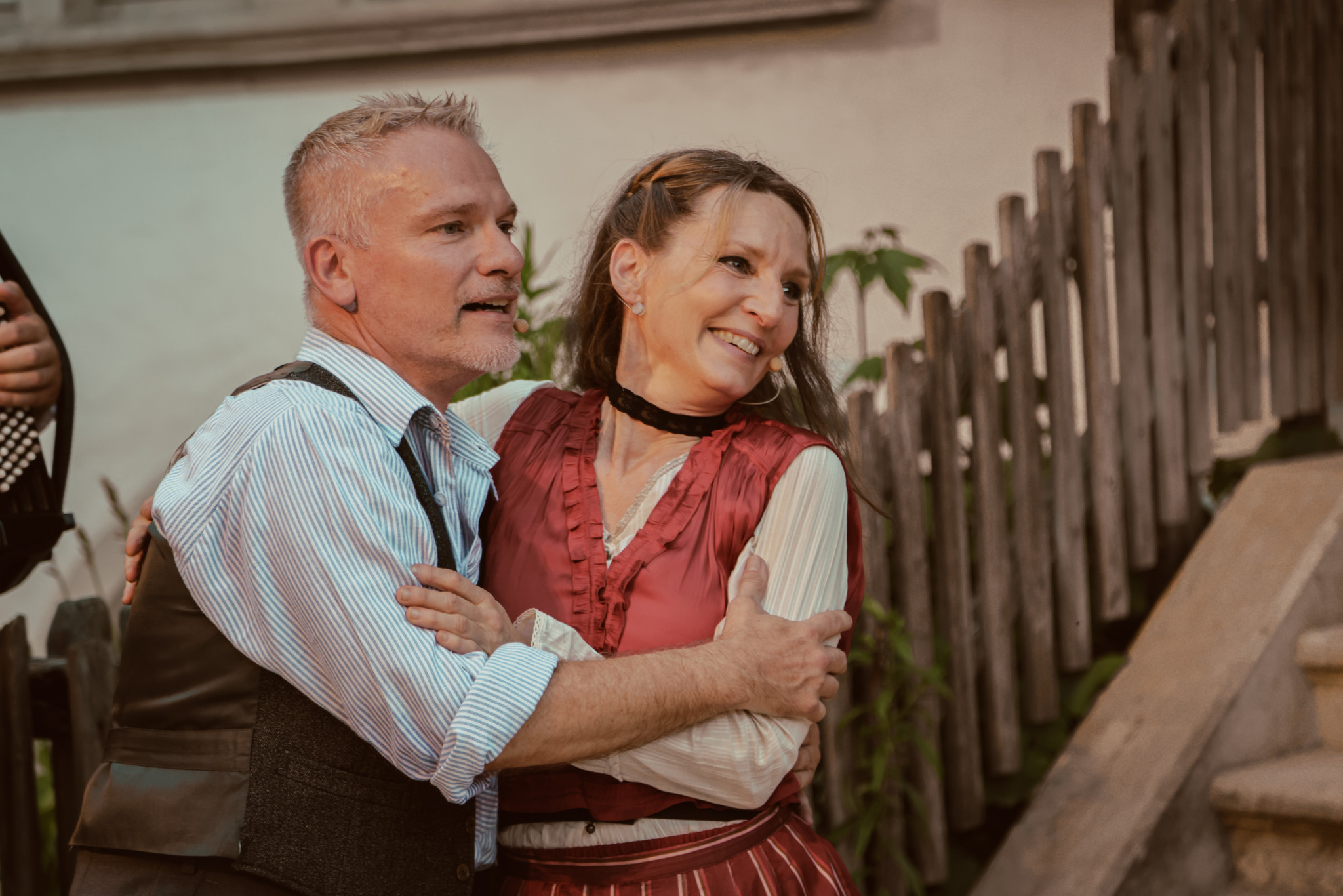 Ein Mann und eine Frau in traditioneller Kleidung lächeln und umarmen sich, während sie nebeneinander stehen. Im Hintergrund ist ein Holzzaun zu sehen.