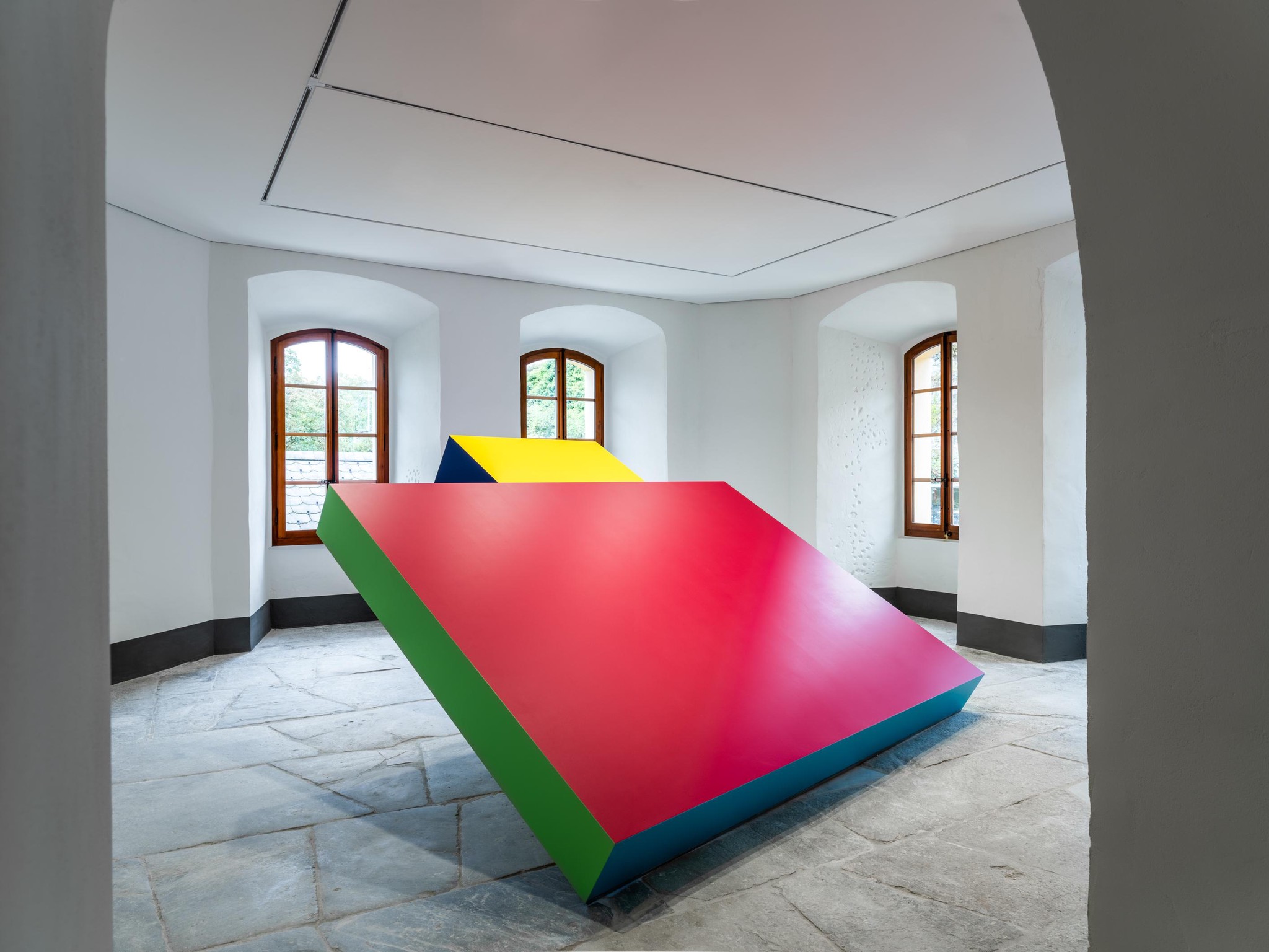 Sculpture géométrique aux couleurs vives rouge, jaune, bleu et vert dans une salle lumineuse avec des murs blancs et des fenêtres cintrées.