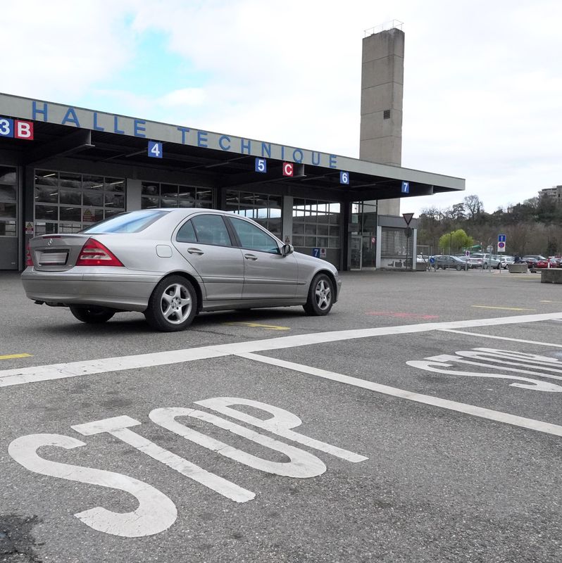 Voiture argentée devant l’office cantonal des véhicules à Carouge, avec un marquage ’STOP’ sur la route.
