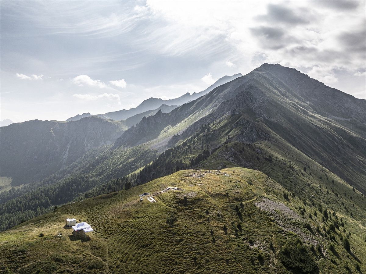 Paysage montagneux avec une chaîne de montagnes verdoyantes sous un ciel légèrement nuageux. Une petite structure blanche est visible sur la colline au premier plan.