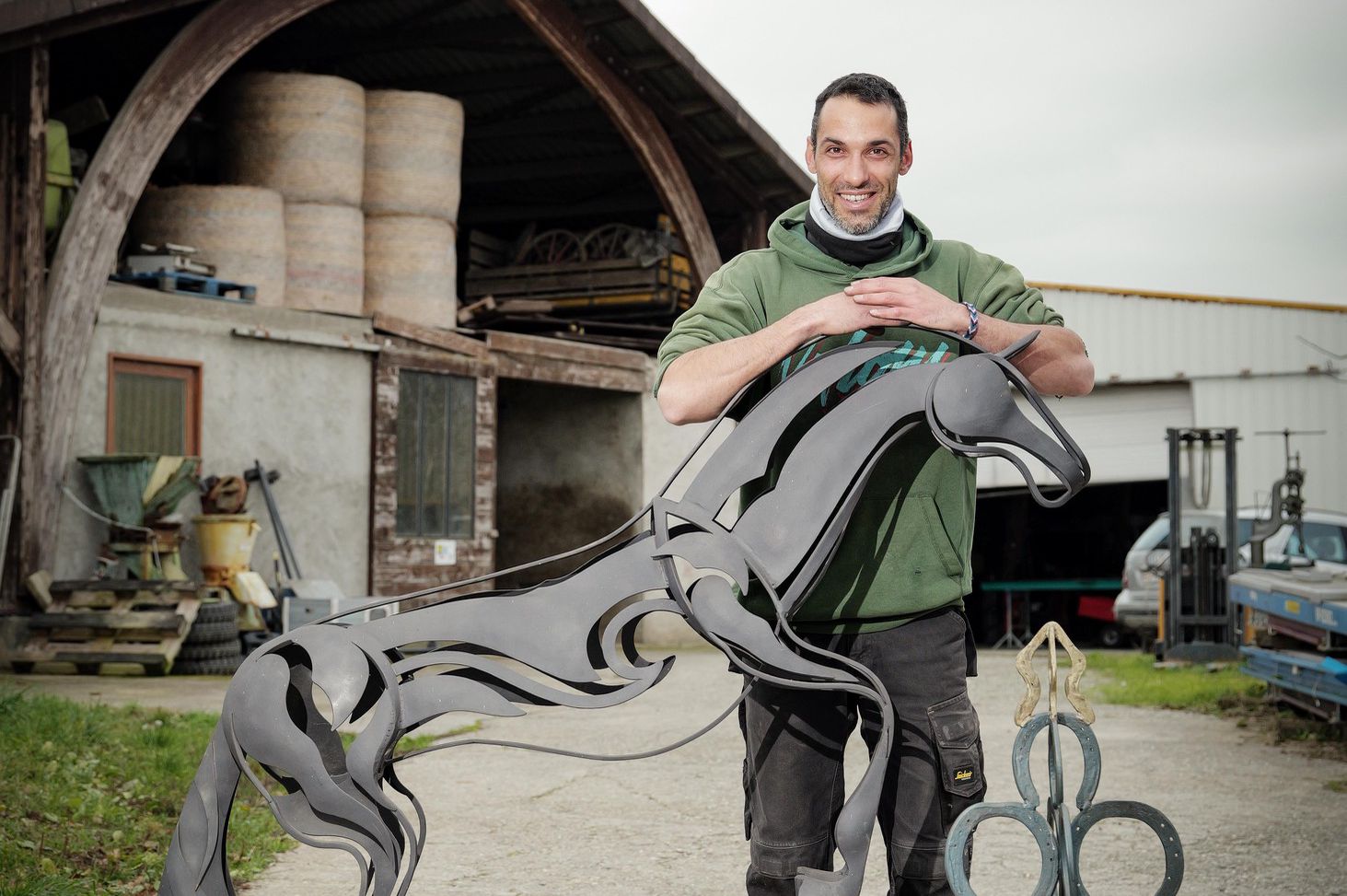 David Ripert, maréchal ferrant et artiste-sculpteur à Avully, pose avec une sculpture en métal représentant un cheval et un empilage de fers à cheval.