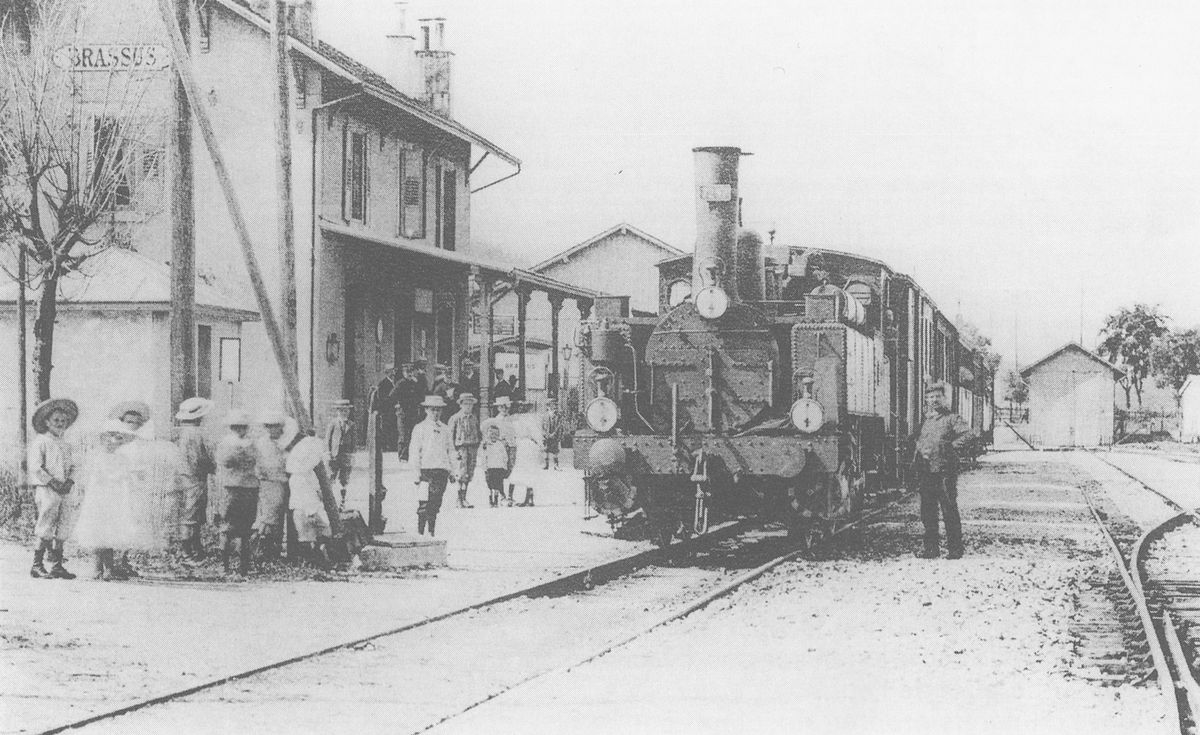 Gare de Le Brassus avec un train à vapeur, entouré de plusieurs personnes dont des enfants, en attente sur le quai.