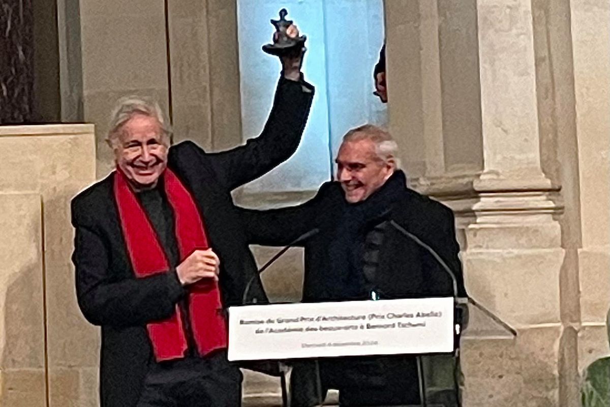 Deux hommes souriants lors d’une cérémonie de remise de prix, l’un d’eux tenant un trophée en l’air. Une pancarte sur le pupitre mentionne un événement de récompense à l’Académie des beaux-arts.