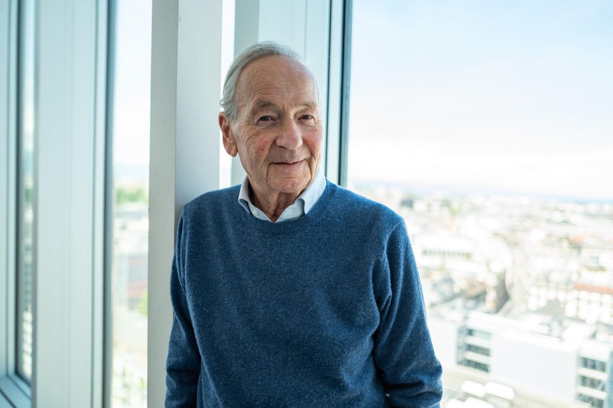Un homme âgé en pull bleu pose près d'une fenêtre avec une vue sur la ville en arrière-plan.