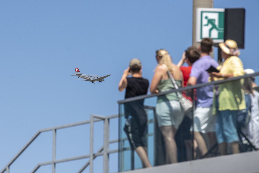 Un groupe de personnes sur une plate-forme d'observation photographient un avion en vol dans un ciel bleu.
