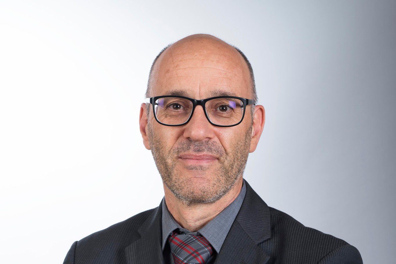 Homme en costume sombre avec lunettes et cravate, bras croisés, sur un fond gris dégradé.