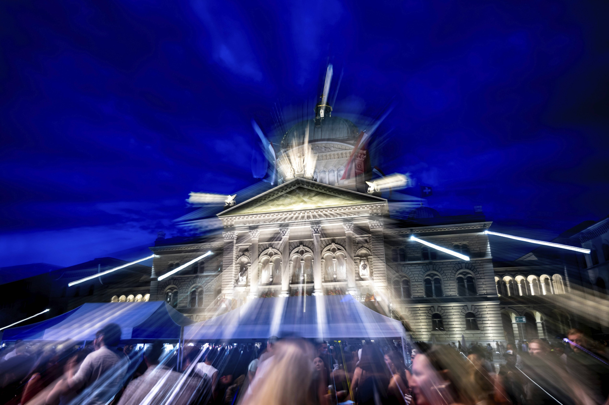 Beleuchtetes Bundeshaus in Bern bei Nacht, umgeben von einer Menschenmenge und einer Statue.