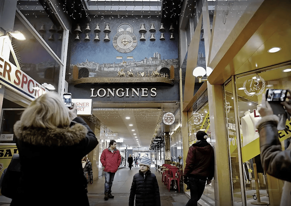 Personnes prennent des photos d’une horloge murale Longines dans une galerie commerçante, avec des boutiques de chaque côté.