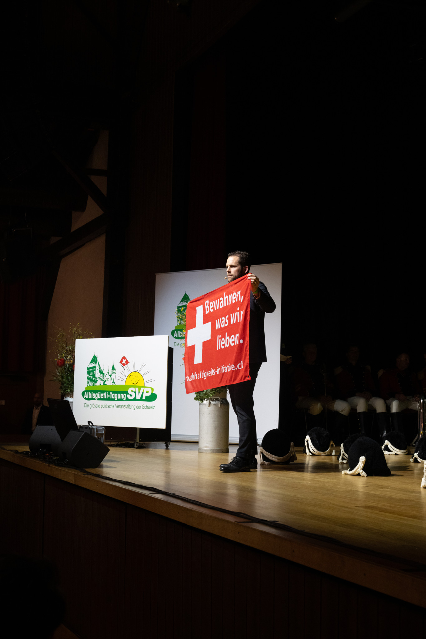 Ein Redner hält ein rotes Plakat mit der Aufschrift ’Bewahren, was wir lieben’ und einem Schweizer Kreuz auf der Bühne der Albisgüetlitagung 2026. Im Hintergrund ist ein Banner der SVP sichtbar.