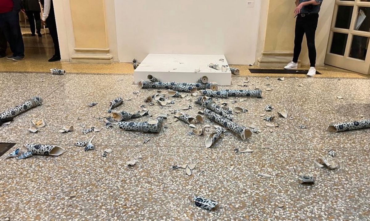 Scène montrant des morceaux de céramique brisée dispersés sur le sol, devant un piédestal blanc dans une galerie d'art.