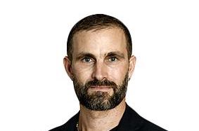 Un homme portant une barbe et un costume sombre pose devant un fond blanc.