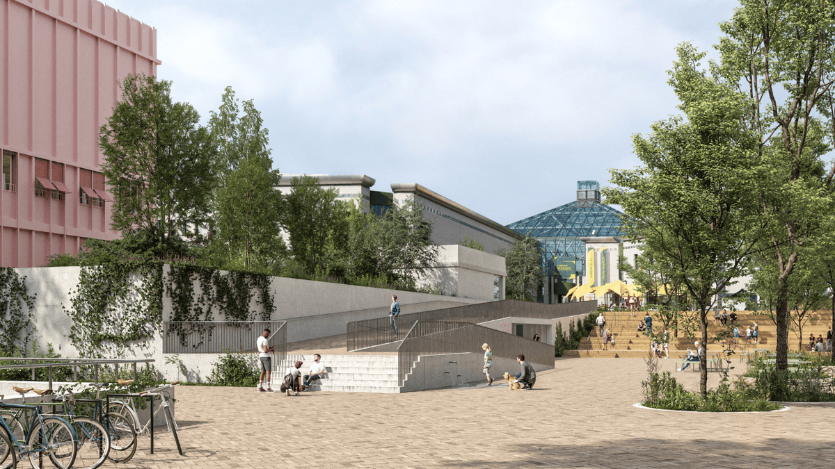 Vue d’une place pavée avec un bâtiment rose et une structure en verre en arrière-plan, entourée d’arbres et de cyclistes.