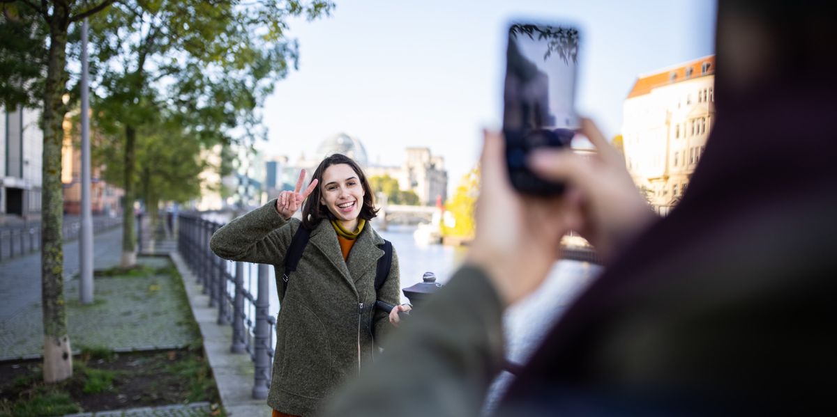 Touriste féminine faisant le signe de la paix devant son petit ami qui la photographie avec un téléphone portable sur une promenade.