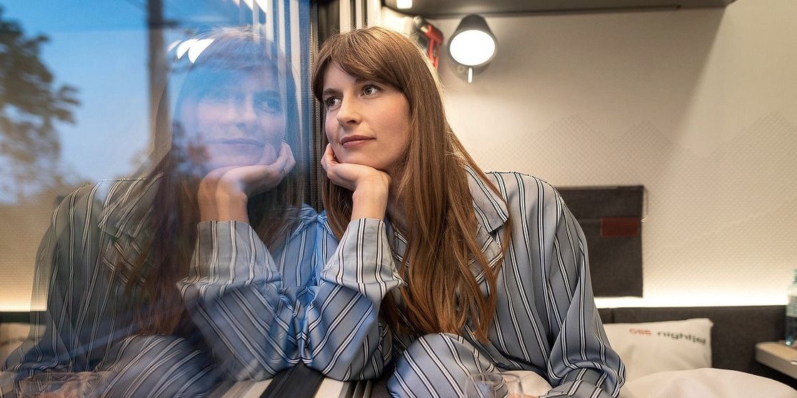 Frau in Pyjama sitzt nachdenklich am Zugfenster, ihr Spiegelbild ist im Glas zu sehen