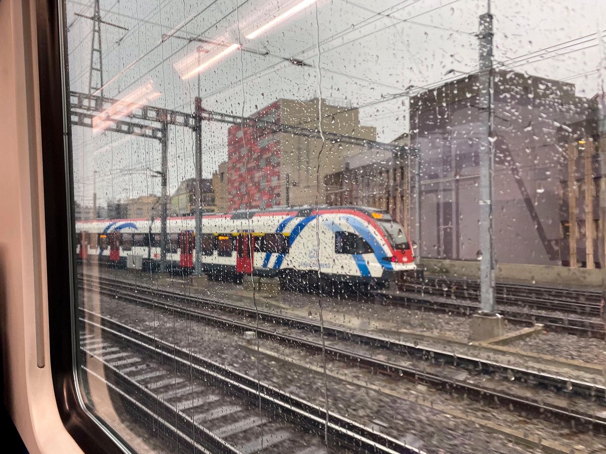 Leman Express à la gare CFF CEVA de Cornavin à Genève sous la pluie, montrant les rails et les bâtiments environnants.