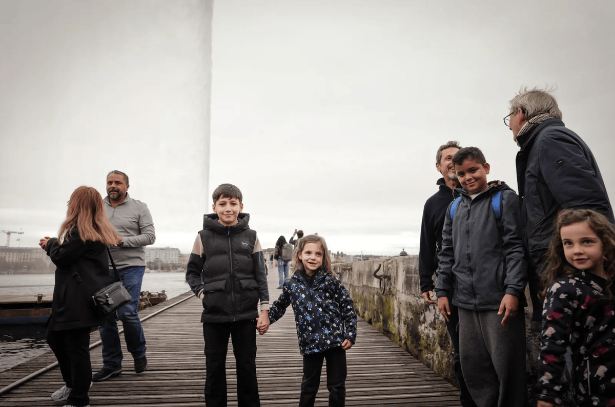 Des enfants souriants se tiennent la main sur un quai en bois à Genève, avec le Jet d'eau en arrière-plan et quelques passants.