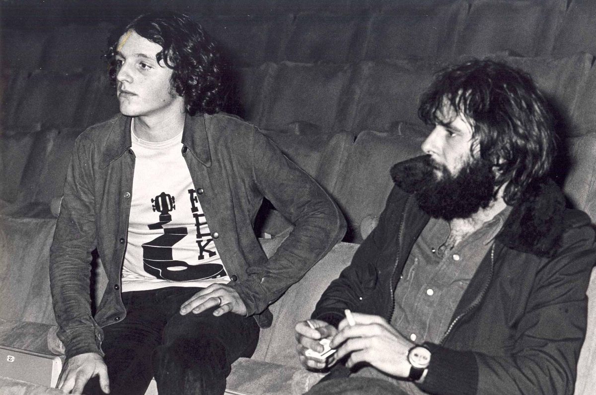 Deux hommes assis dans des sièges de théâtre, l’un portant une veste et un t-shirt imprimé, l’autre avec une barbe et tenant un objet.