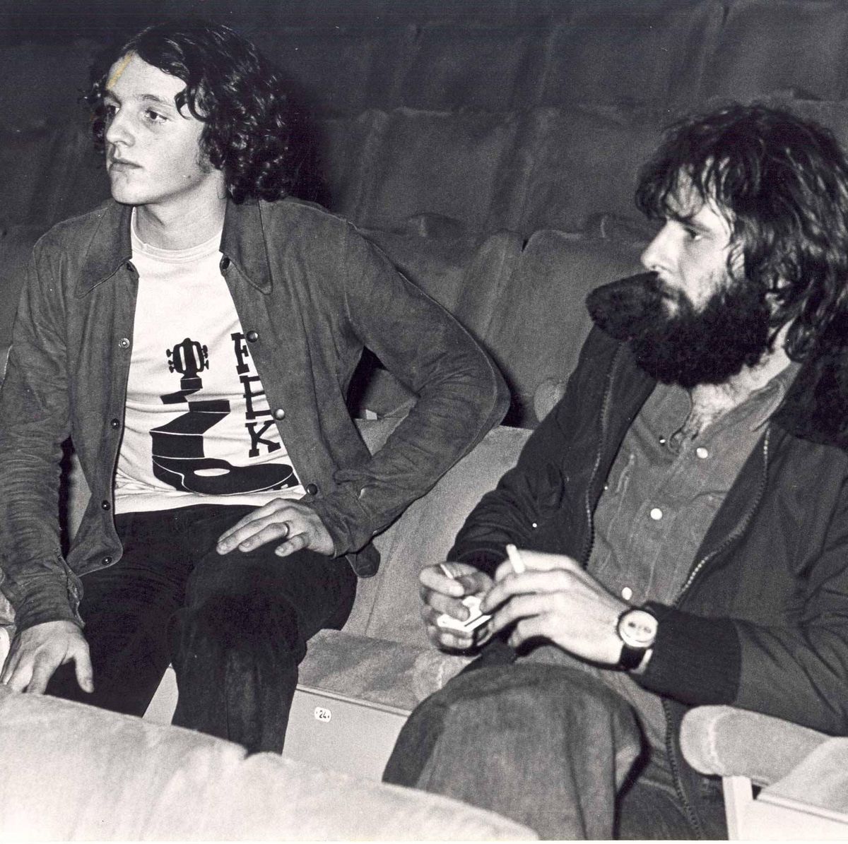 Deux hommes assis dans des sièges de théâtre, l’un portant une veste et un t-shirt imprimé, l’autre avec une barbe et tenant un objet.