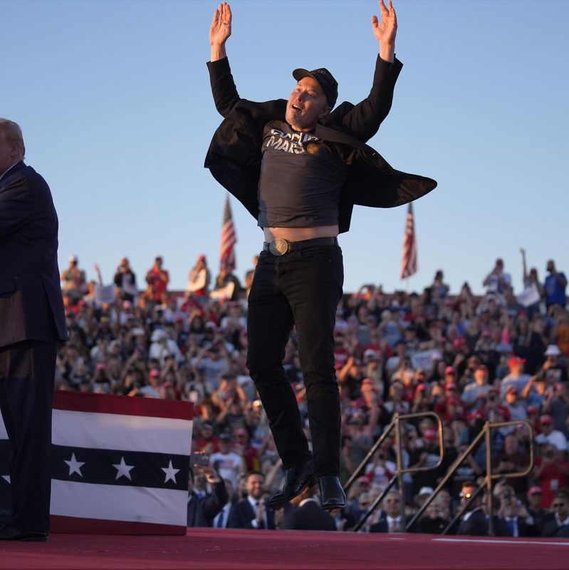 Personne en train de sauter sur scène pendant qu’un orateur se tient au pupitre lors d’un rassemblement en plein air avec une foule en arrière-plan.