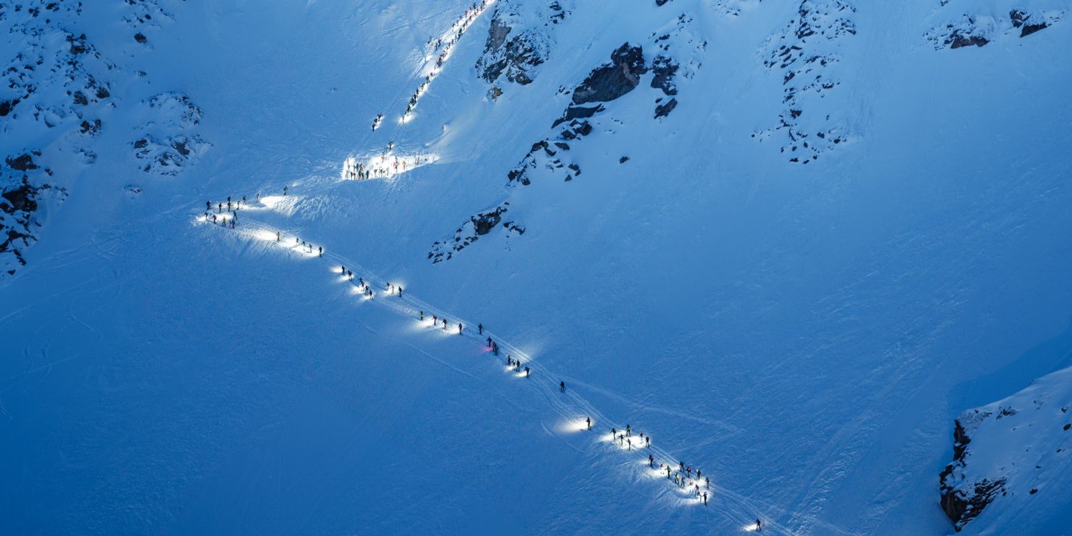 Une longue file de participants à la Patrouille des Glaciers avec des lampes frontales escaladant une montagne enneigée sous un ciel bleu sombre.